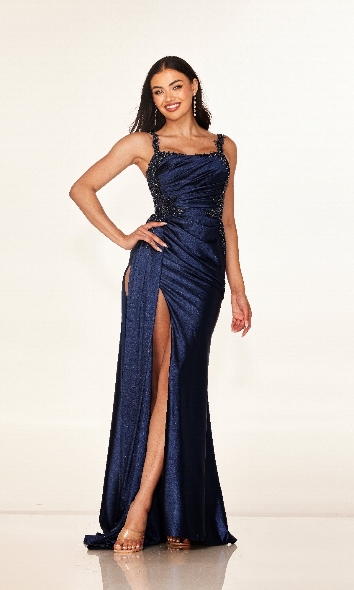 A woman smiles against a plain background wearing the Cecilia Couture 2612 long prom dress, a navy blue, form-fitting gown with beaded straps, side embellishments, and a high leg slit.